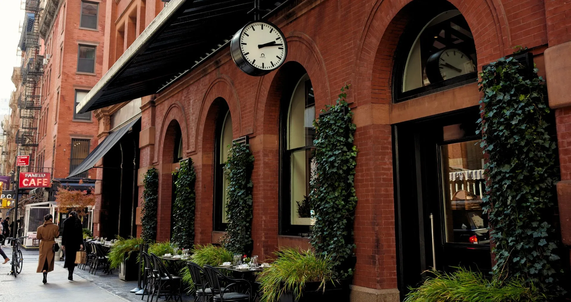 Outdoor seating area with tables and plants along a brick building with arched windows and a clock.