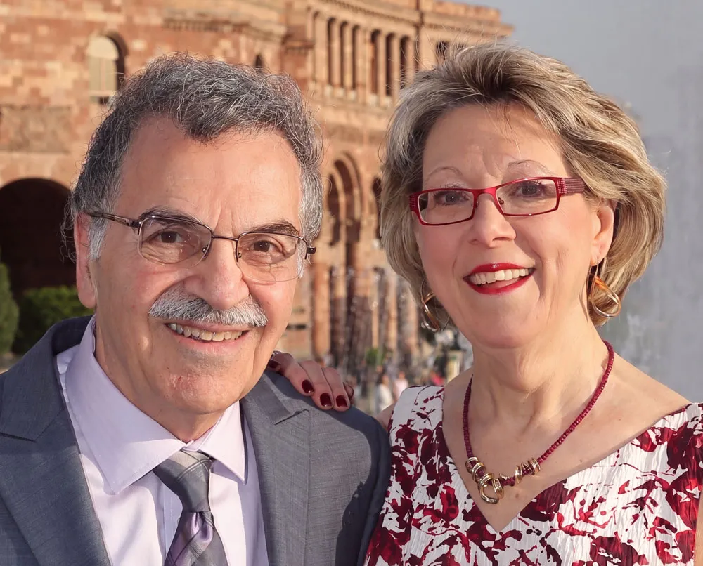 Smiling elderly man and woman posing outdoors with historic brick building in background.
