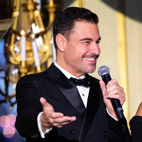 Man in a black tuxedo smiling and speaking into a microphone with a golden chandelier in the background.