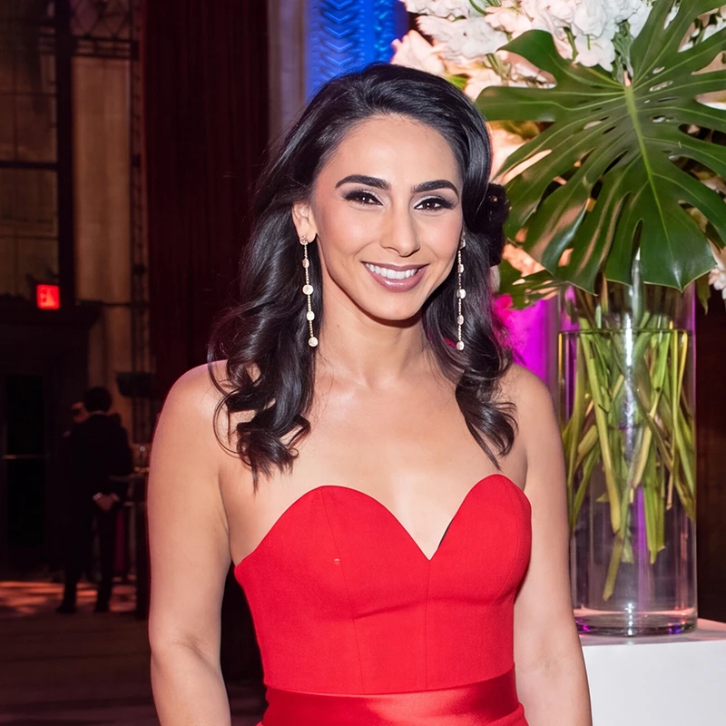 Smiling woman with dark hair wearing a red strapless dress and long earrings at an indoor event.