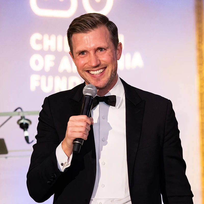 Man in a tuxedo holding a microphone and smiling at an indoor event.