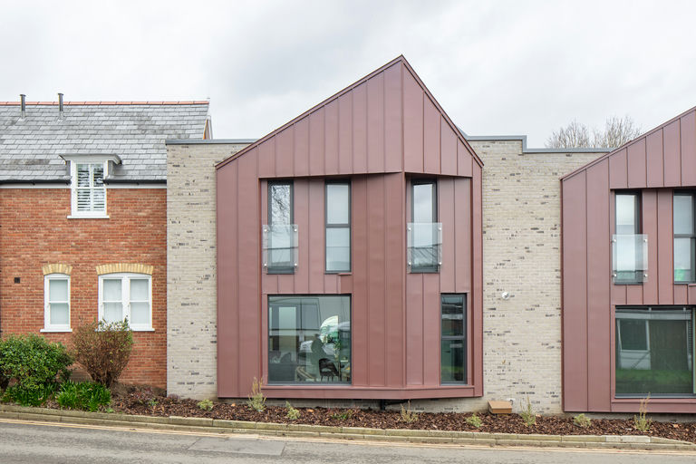 A modern architectural design featuring two adjoining buildings along a street. The building on the right has a contemporary style with vertical red-brown metal cladding and large rectangular windows, contrasting with the traditional red brick house on the left that features smaller, classic white-framed windows and a pitched roof. The transition emphasizes the blend of old and new architectural styles.