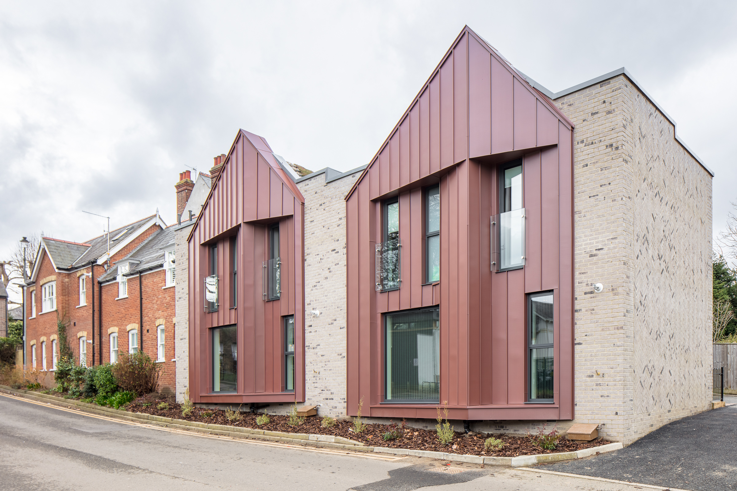 Two modern gabled houses with reddish-brown metal cladding next to a traditional brick house on a cloudy day. The modern houses feature large windows and a minimalist design, contrasting with the older brick building.