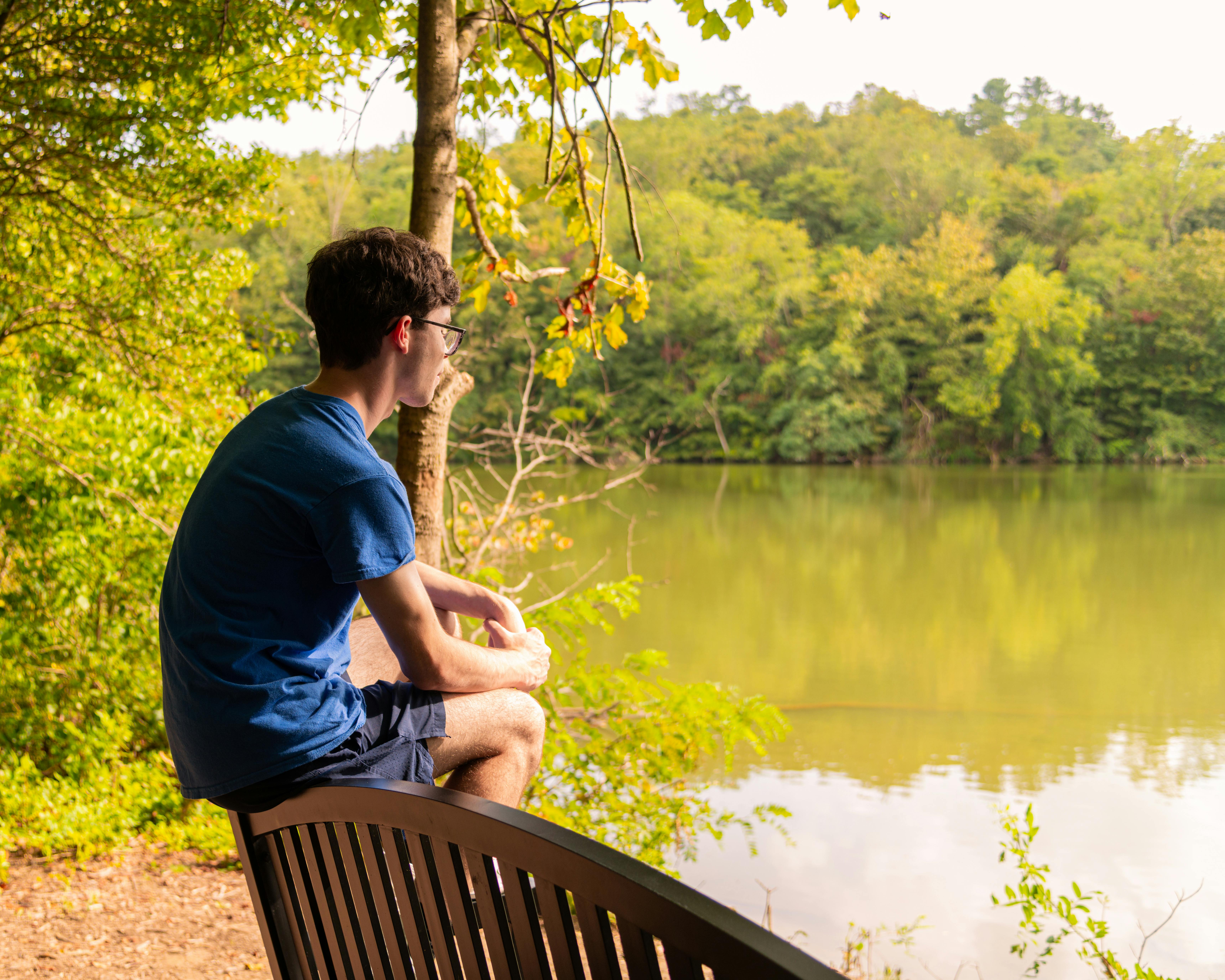 A young man wearing a blue shirt and glasses sits on the back of a bench, looking out over a calm lake surrounded by lush green trees on a bright day.