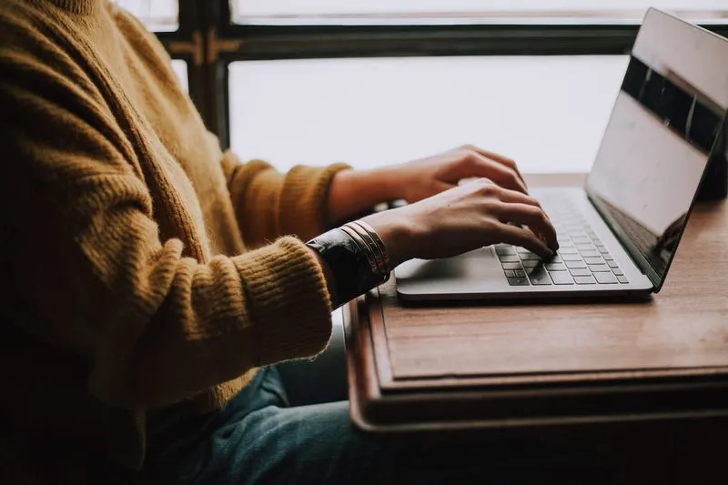 Person in a mustard sweater typing on a laptop at a wooden desk next to a window