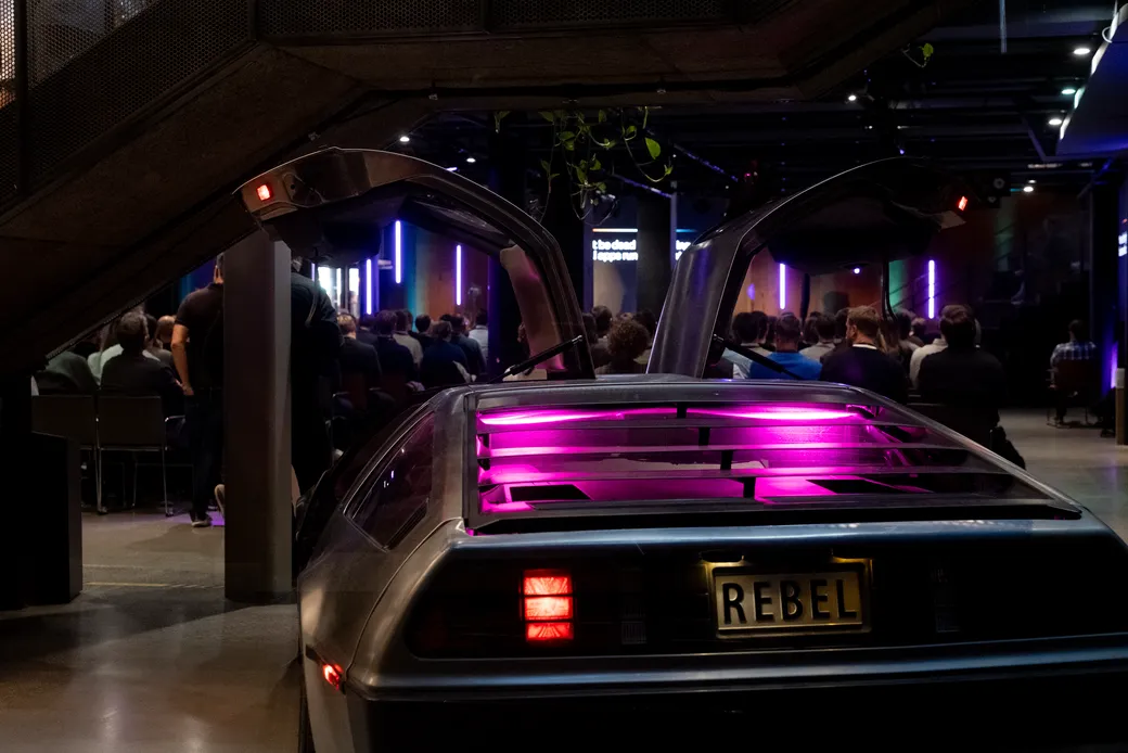 A DeLorean with its gullwing doors open and a REBEL number plate, parked behind the seated Developalooza audience at Rebel in Oslo