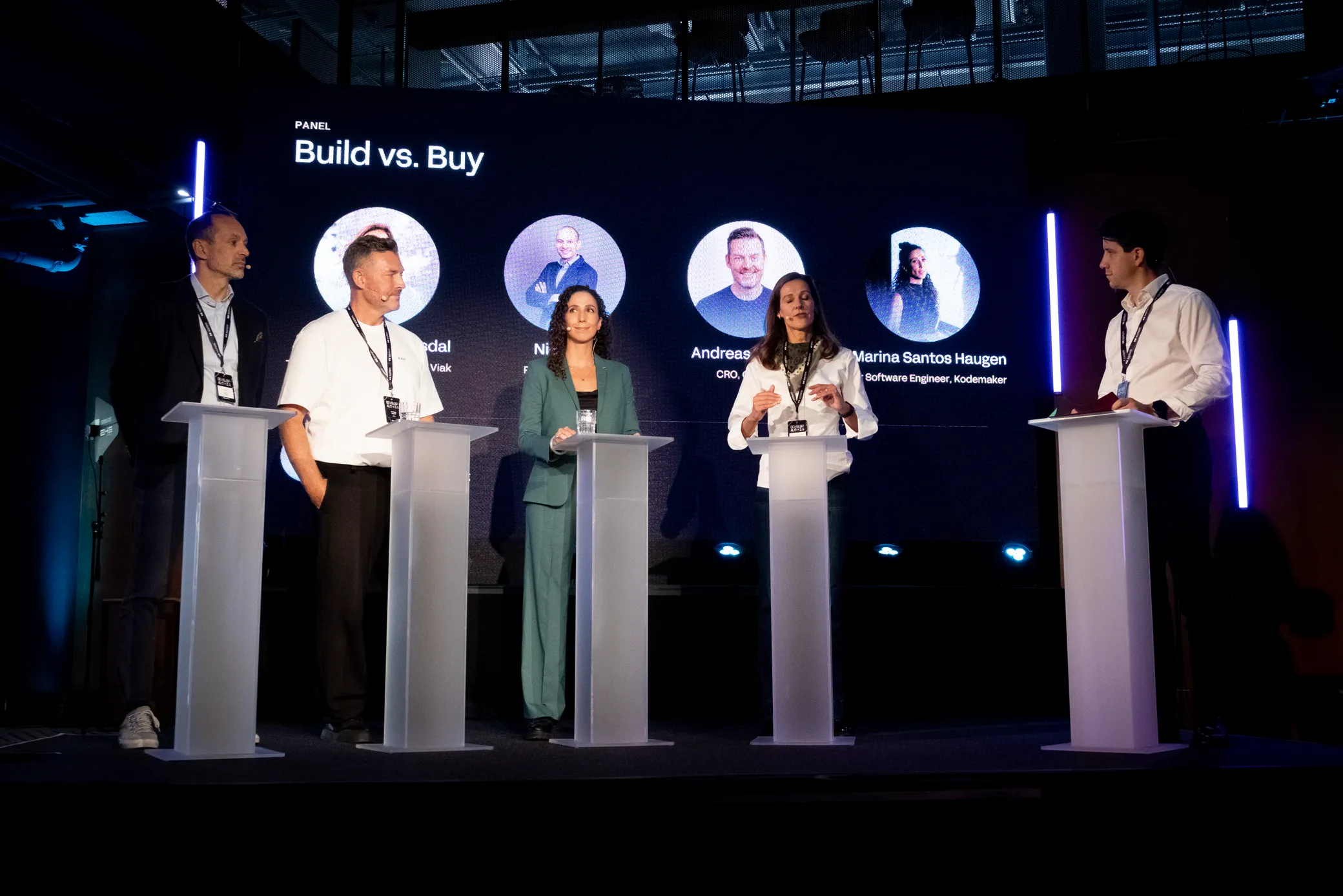 The Build vs Buy panel on stage at Rebel, with the session title and speaker headshots on the screen behind them