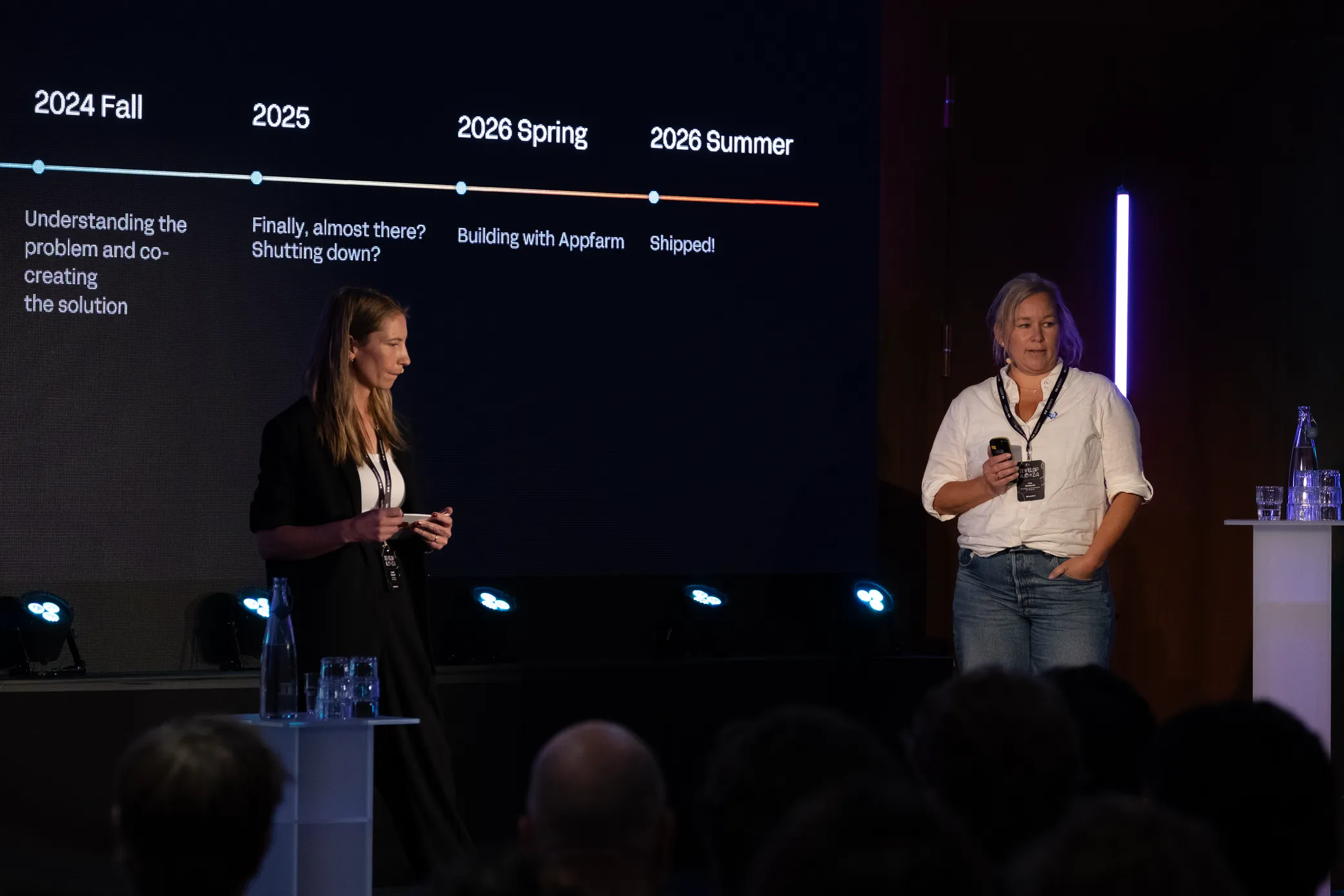 Two presenters on stage in front of a project timeline running from understanding the problem in 2024 to shipped in summer 2026