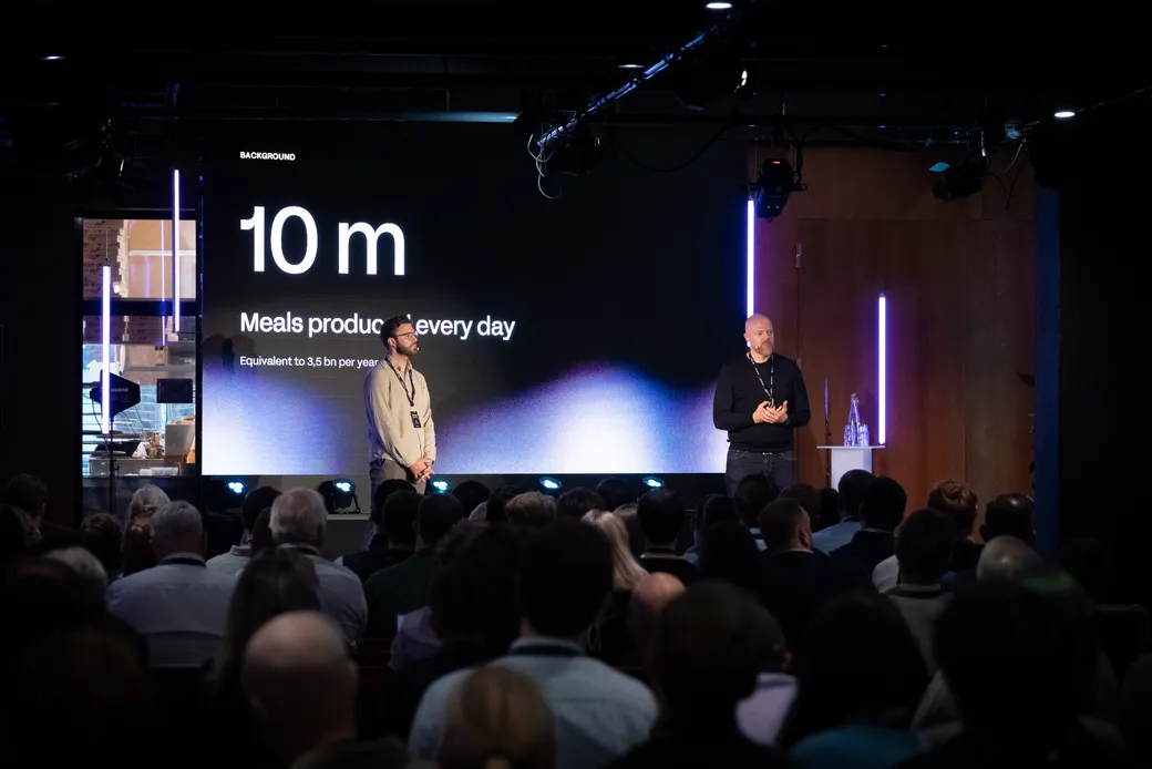 Two presenters on stage in front of a slide reading 10 m meals produced every day, equivalent to 3.5 billion per year