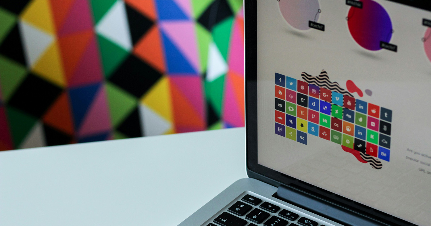 Laptop with colorful graphic design software open on screen, with a bright geometric wall pattern in the background.