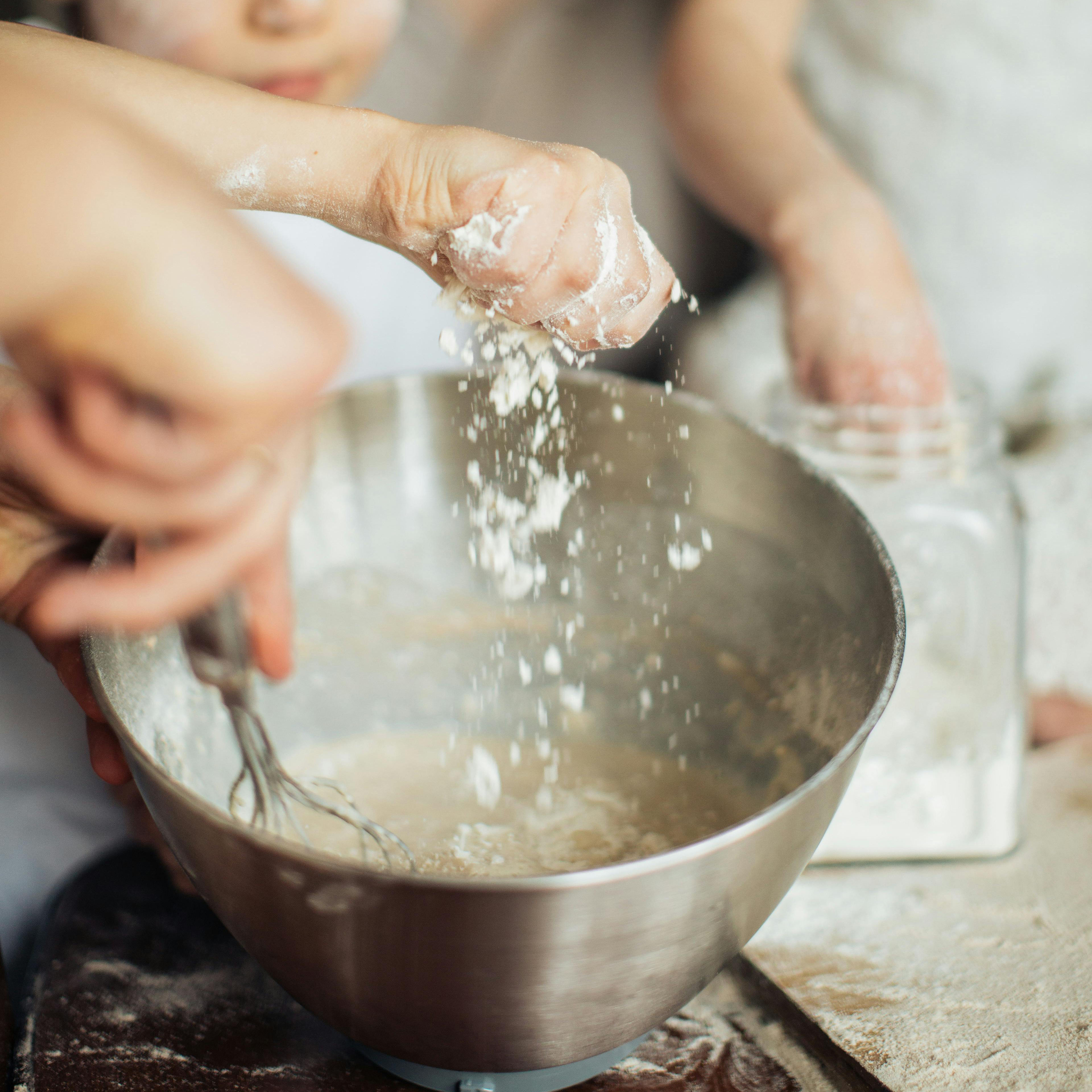 A photo of two kid's hands dropping flour into a mixing bowl. An adult is holding the bowl and mixing with a whisk.
