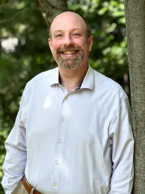 A bald smiling man smiles leaning against a tree