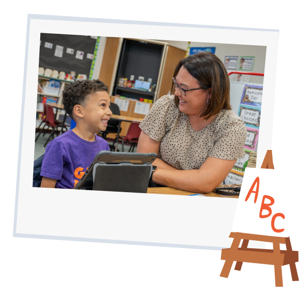 Young boy and teacher smiling in classroom setting while using Khan Kids on tablet. 