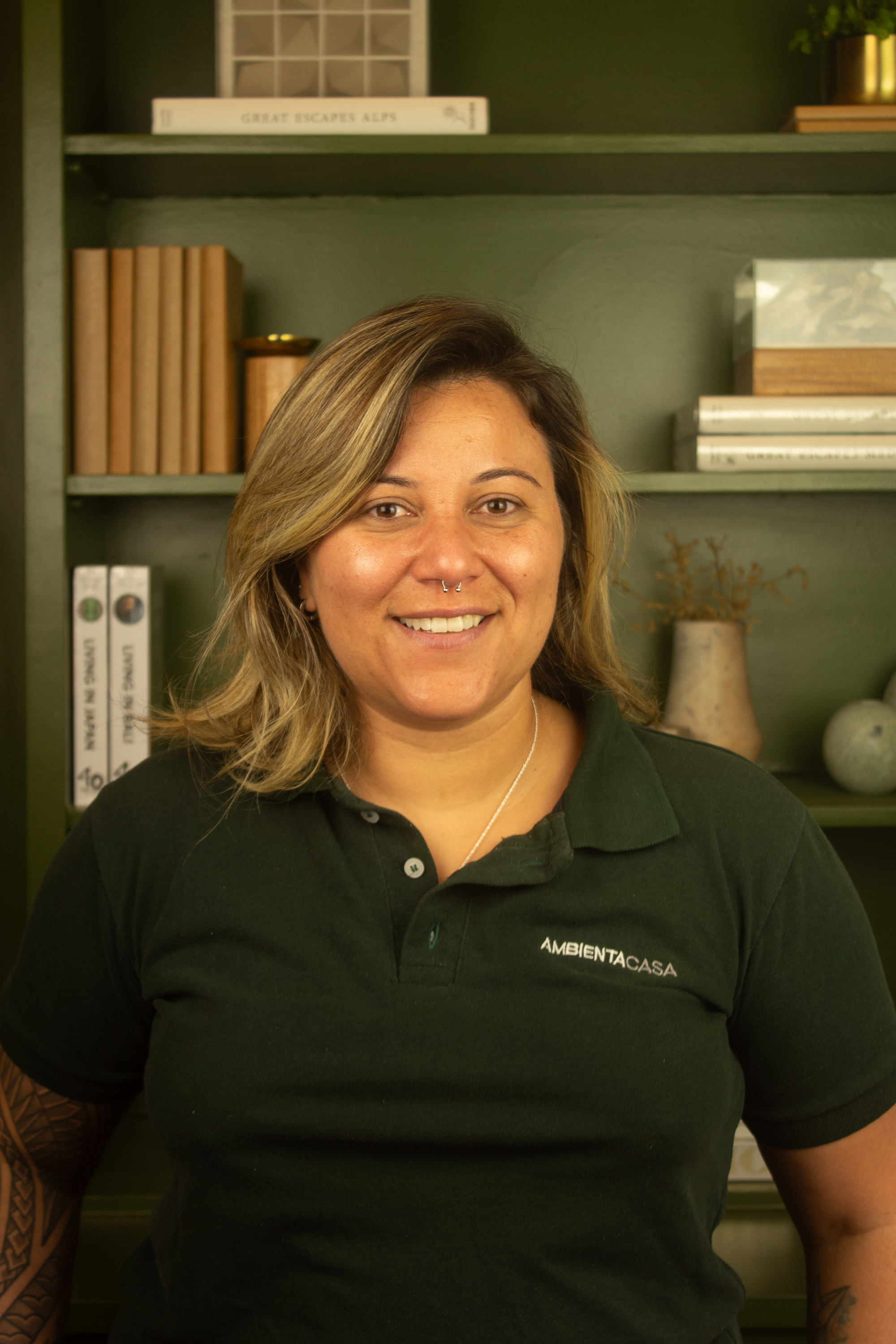 Juliana Dias, colaboradora da Ambienta Casa, com uma camisa polo verde e uma estante com livros e decorações atrás.