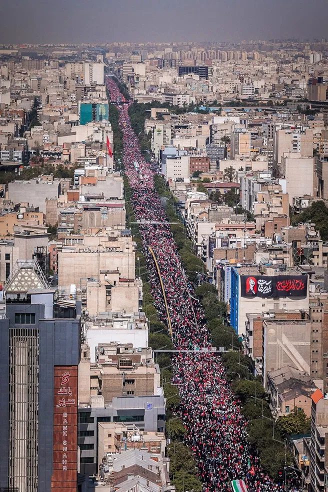 Crowds fill a Tehran avenue beneath red mourning banners and national flags.