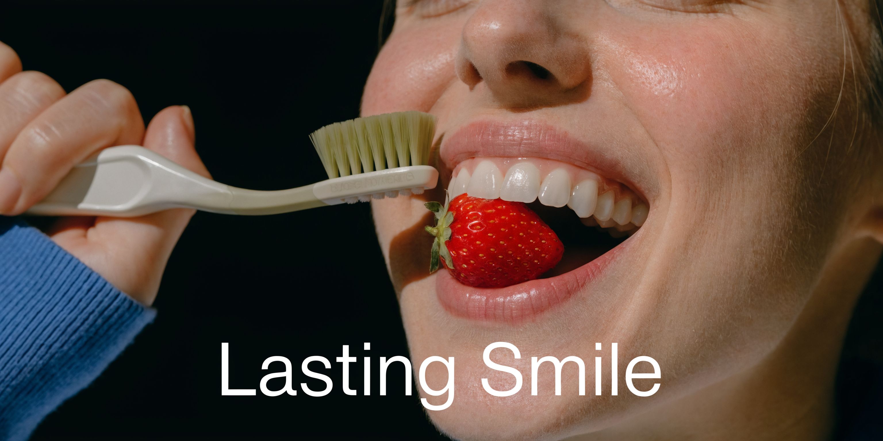 A close-up of a person holding a toothbrush near their teeth while holding a fresh red strawberry.