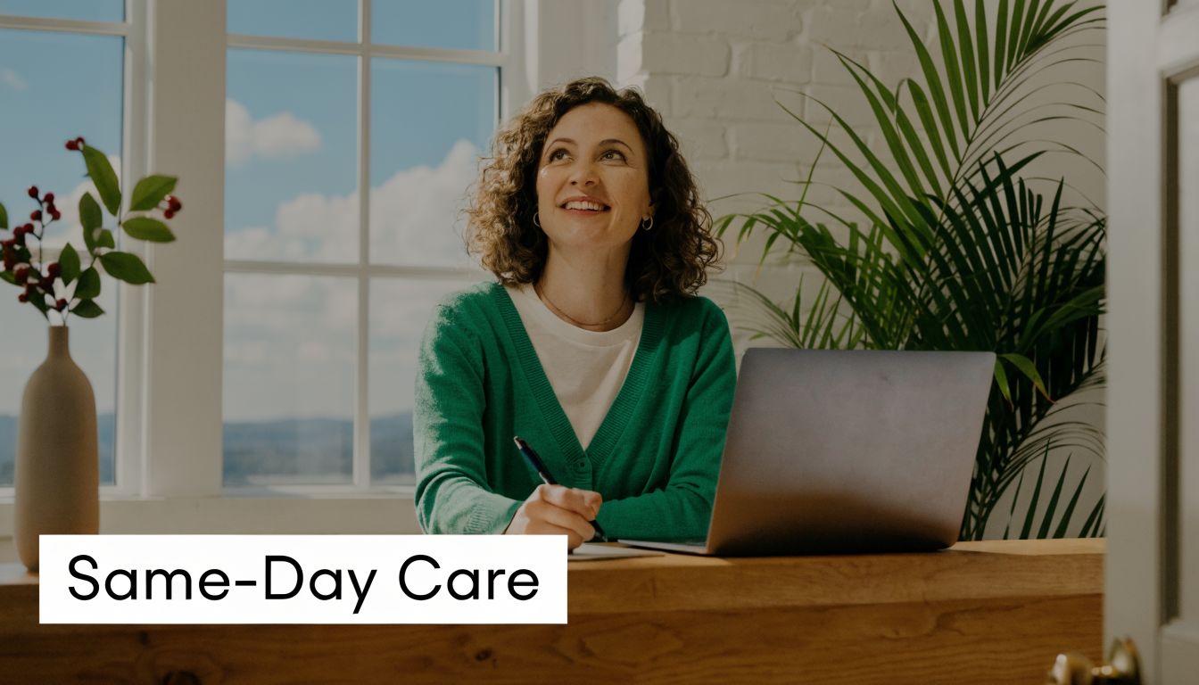 A smiling woman with curly hair sits at a desk working on a laptop near a window.
