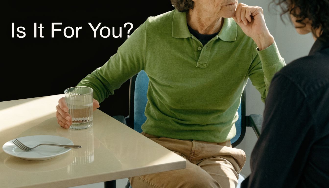 An elderly man in a green shirt holds a glass while sitting at a dining table.