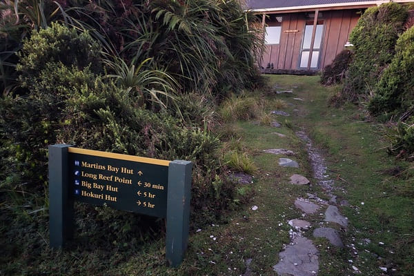 A photo of a New Zealand Department of Conservation Track sign to Martins Bay Hut, with the hut in the background