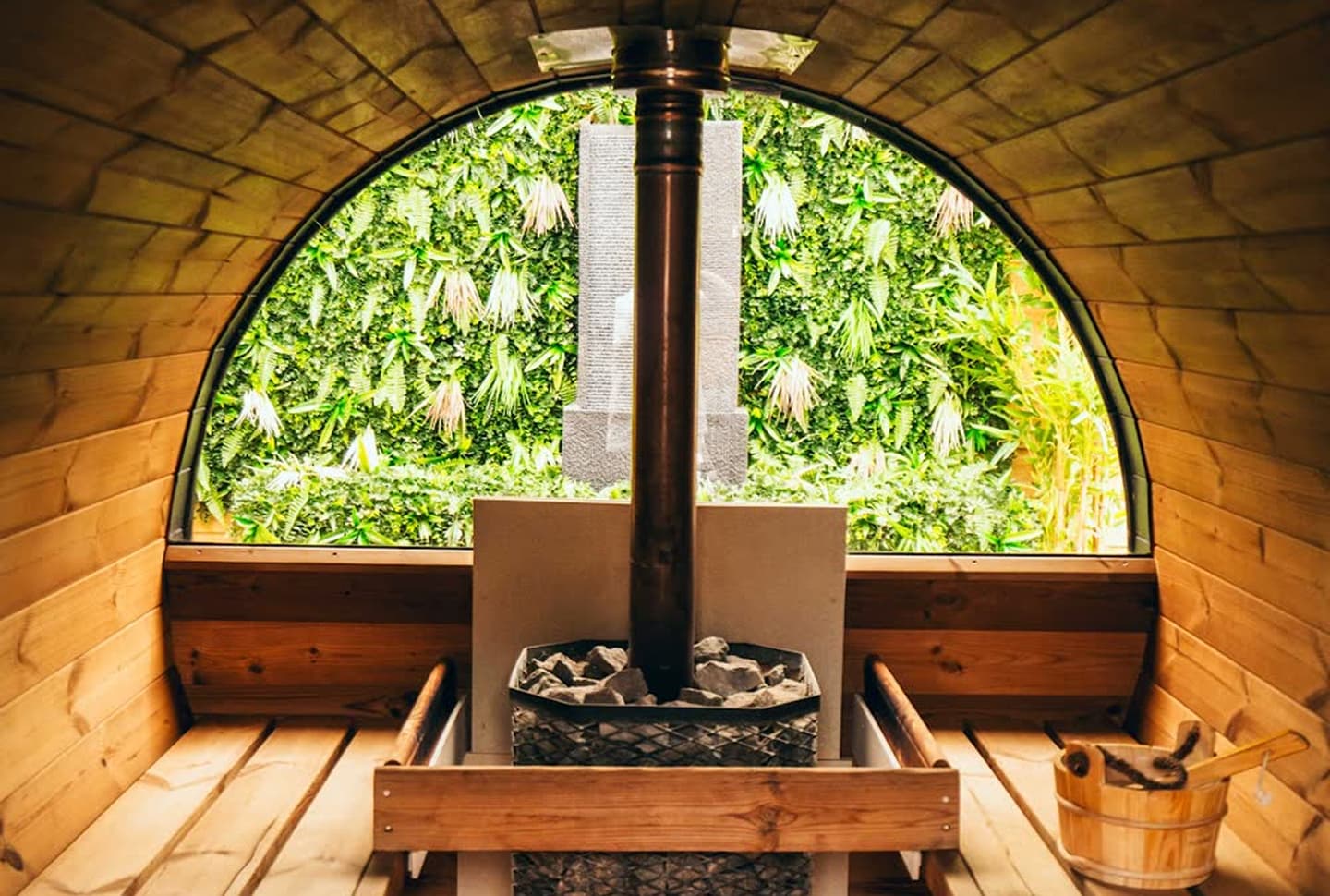 Interior of a wooden barrel-shaped sauna with a central stove filled with sauna rocks, wooden benches on both sides, and a wooden bucket with a ladle on the right; a large arched window shows green leafy plants outside.