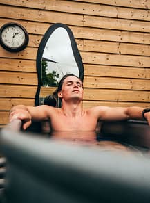 A shirtless man relaxing in an outdoor hot tub with arms resting on the sides, against a wooden slat wall with a clock and a tall mirror.