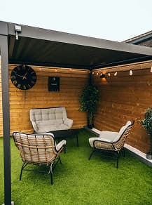 Outdoor seating area with three cushioned chairs around a small round table under a black pergola, artificial green grass, wooden paneled walls with a clock and wall decoration, and string lights above.