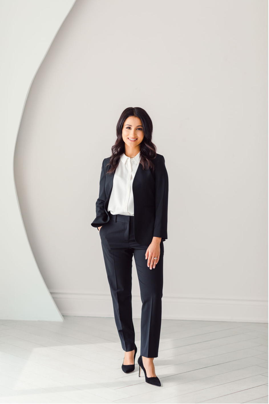Woman in a black suit with a white blouse smiling while standing in a bright room with a light curved wall detail.
