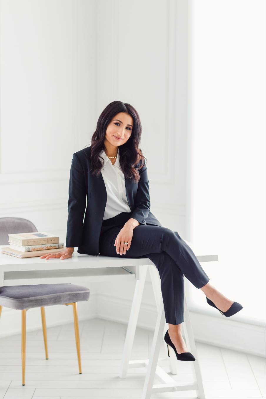 Woman in black suit and white blouse sitting on a white desk in a bright room with books stacked beside her.