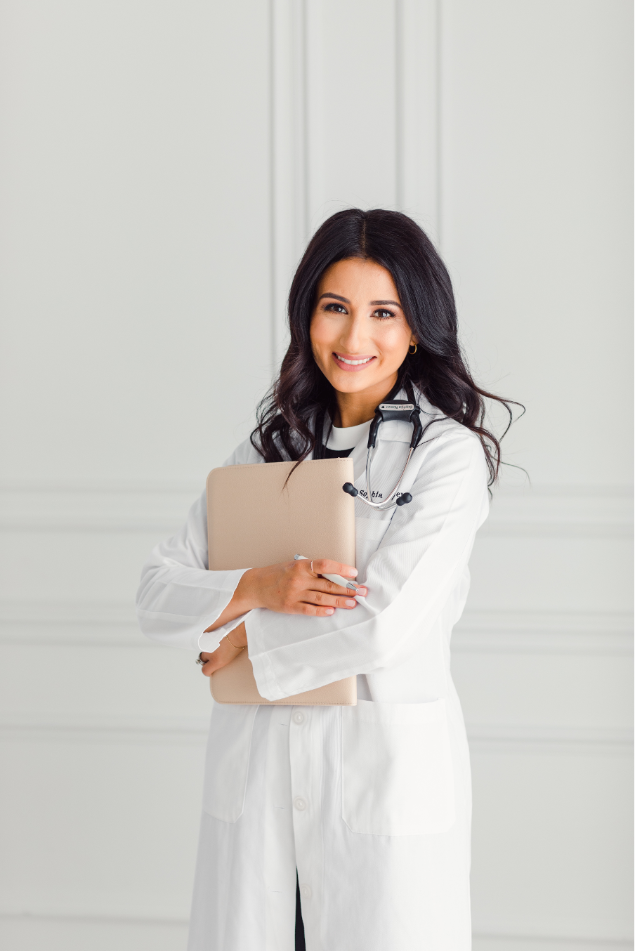 Smiling female doctor with dark hair wearing a white coat and stethoscope, holding a beige folder.