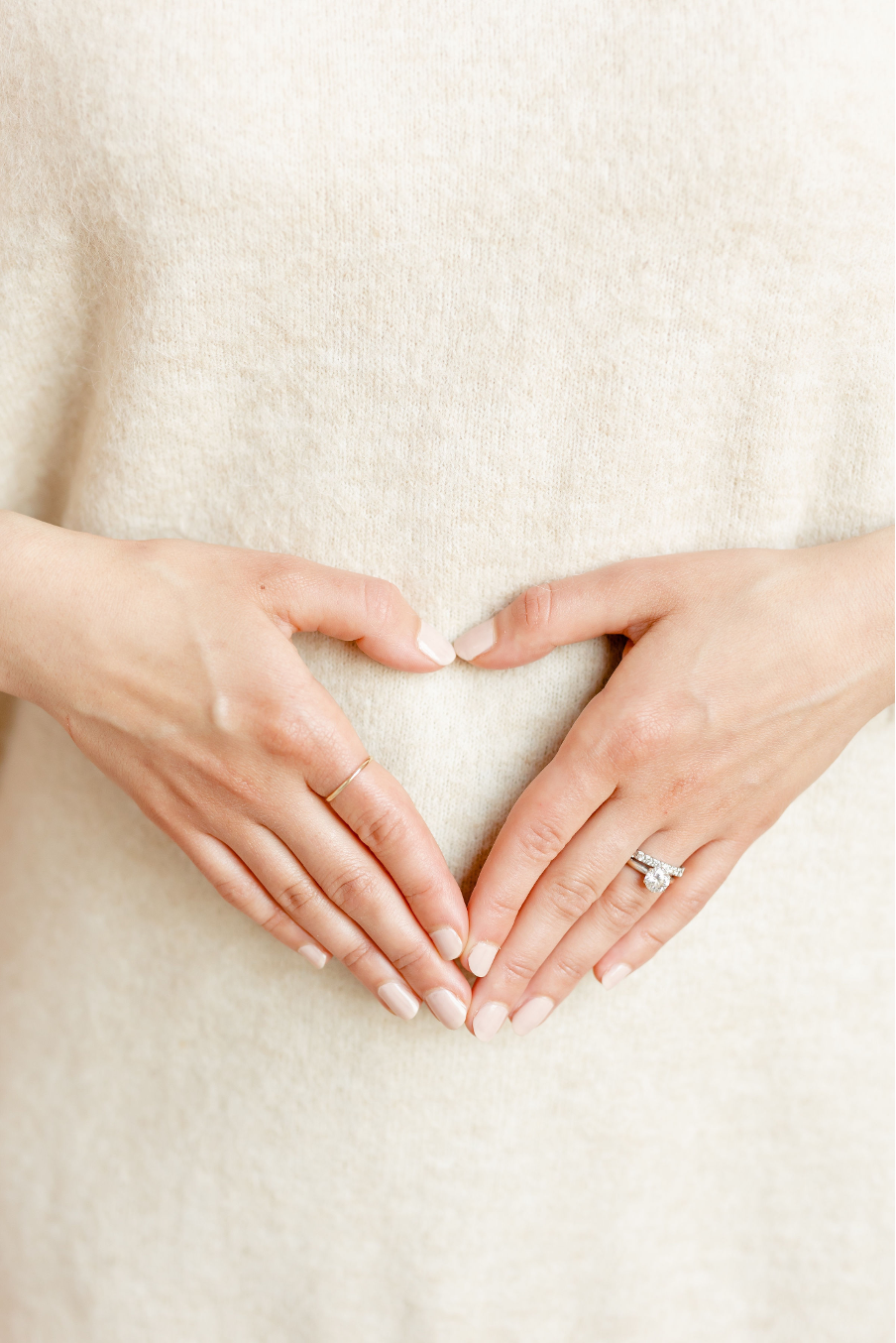 Close-up of hands forming a heart shape over a light beige sweater, with a ring on the right hand.