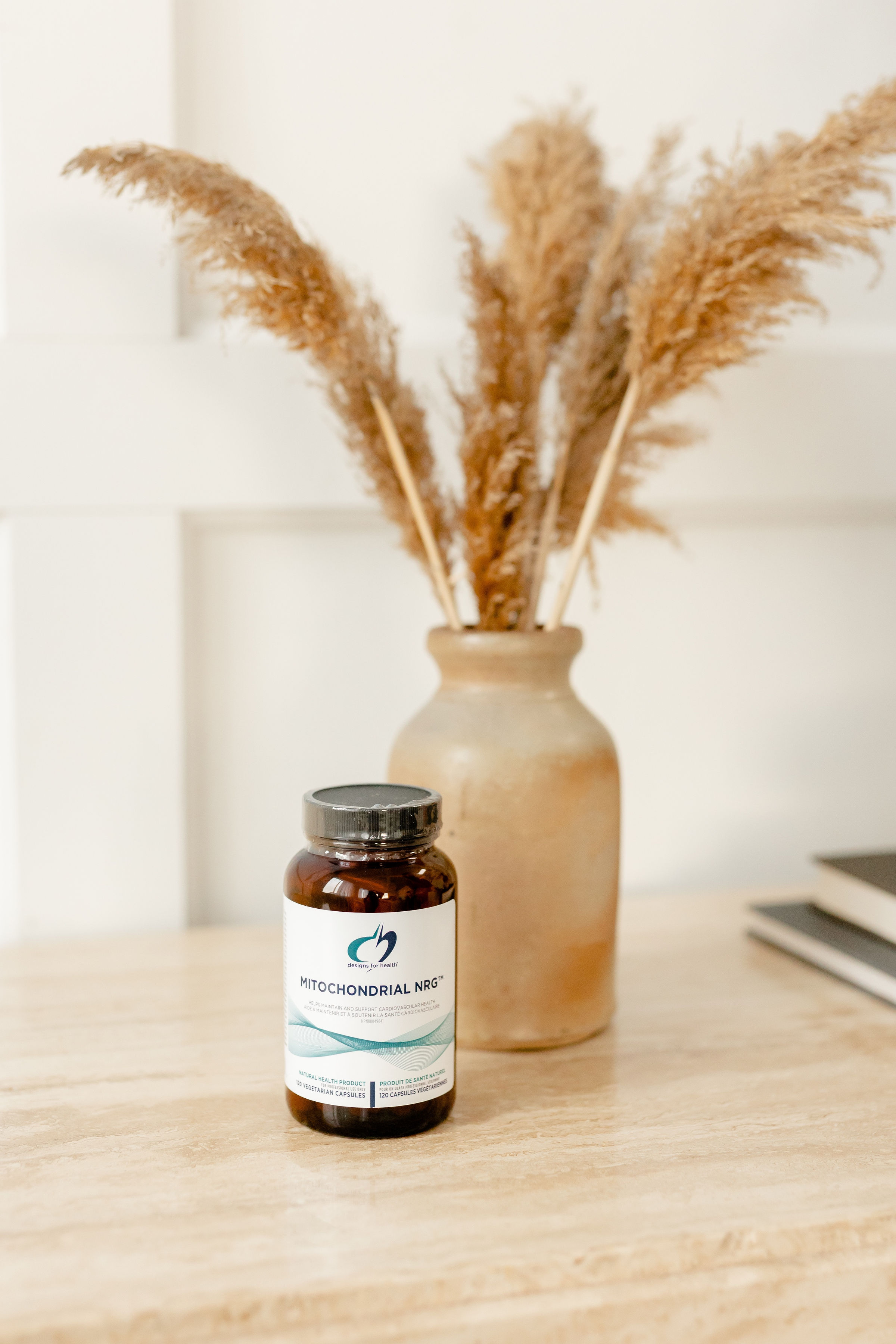 Brown supplement bottle labeled Mitochondrial NRG on a beige table with a ceramic vase holding dried pampas grass in the background.