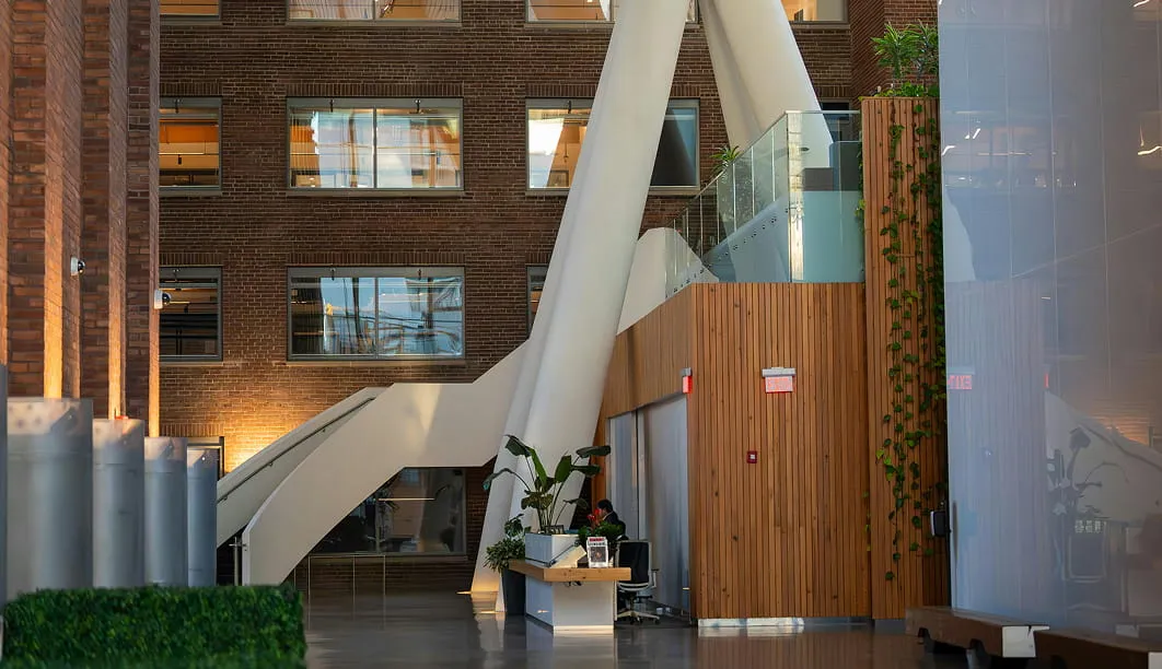 Modern office lobby with large white support beams, brick walls, plants, and glass railings.
