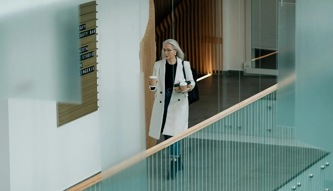 Mature woman in white coat carrying multiple coffee cups while walking in a modern building.