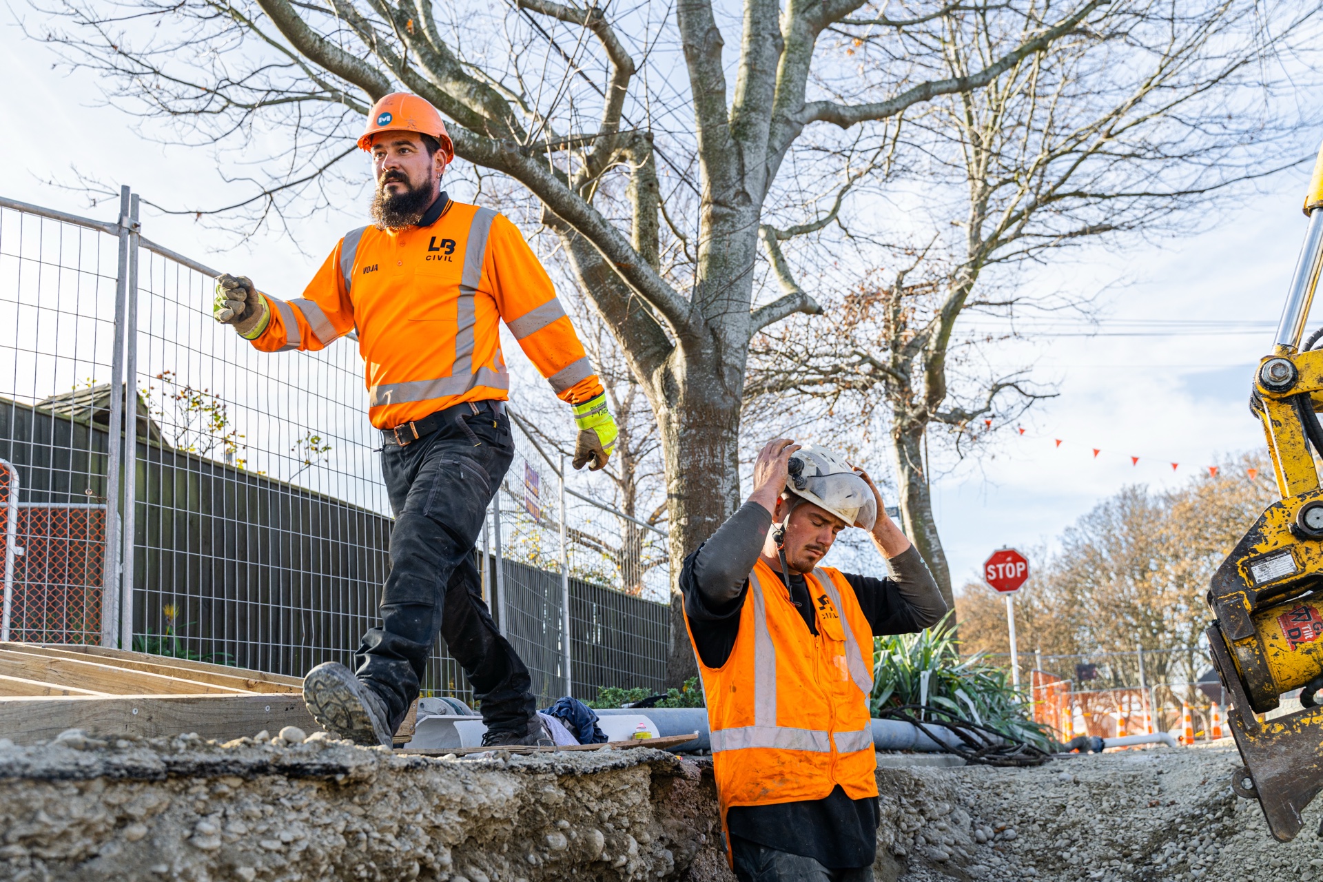 Two workers at the edge of an open trench, one adjusting a hard hat.