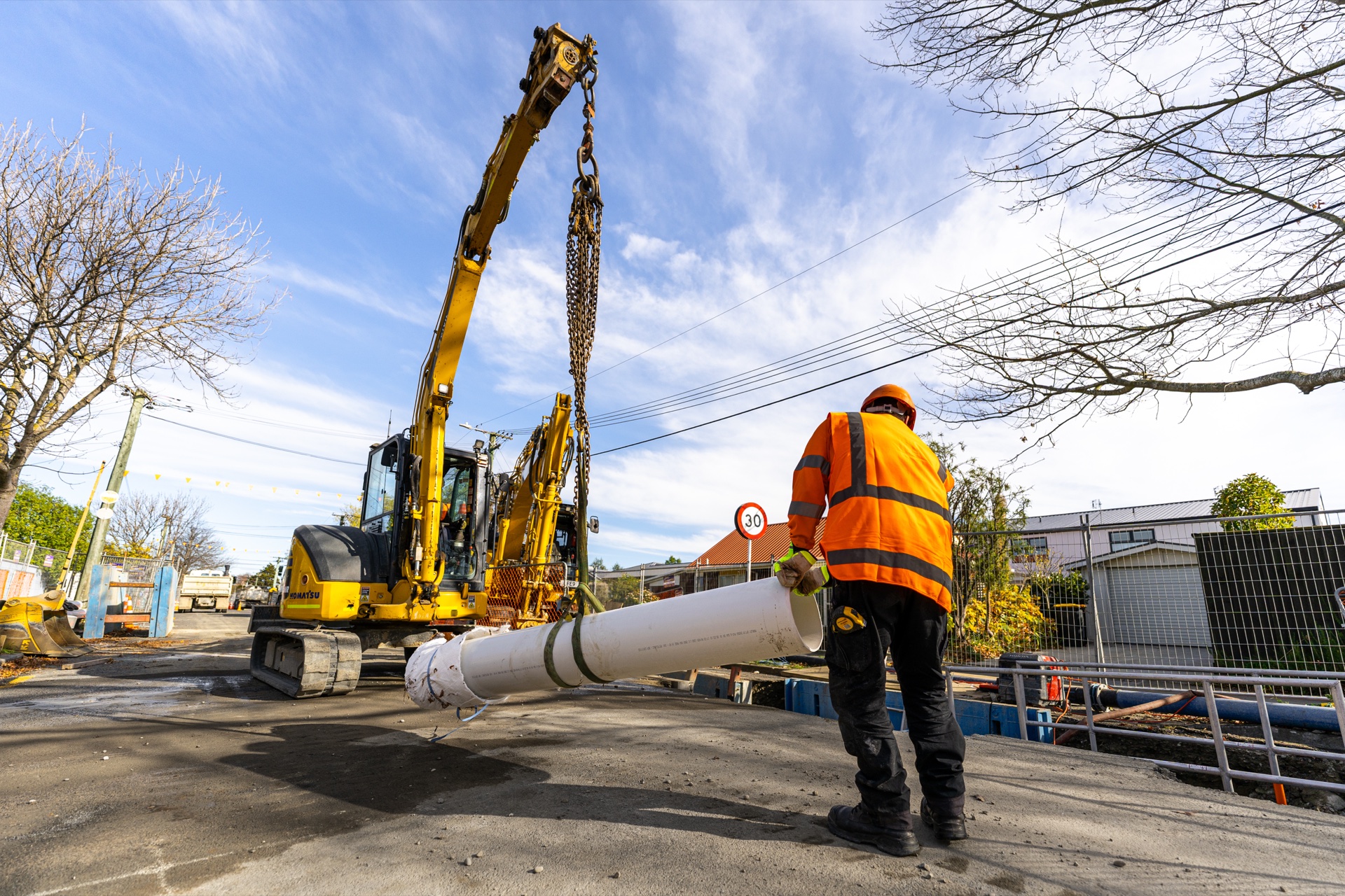 Excavator lifting a long white pipe into position with a worker standing by.