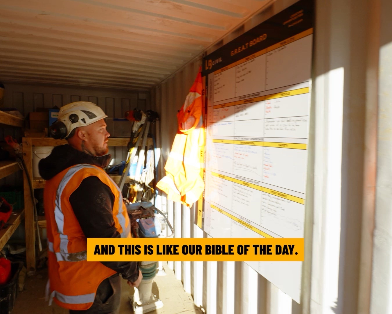 Worker in a site container reading the GREAT board, the crew's daily job planning whiteboard.