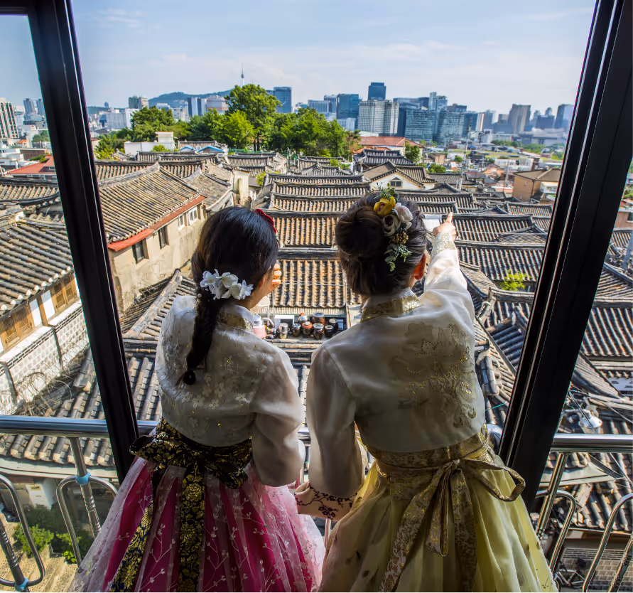 Deux femmes en hanbok observant le village hanok de Bukchon à Séoul depuis un balcon vitré