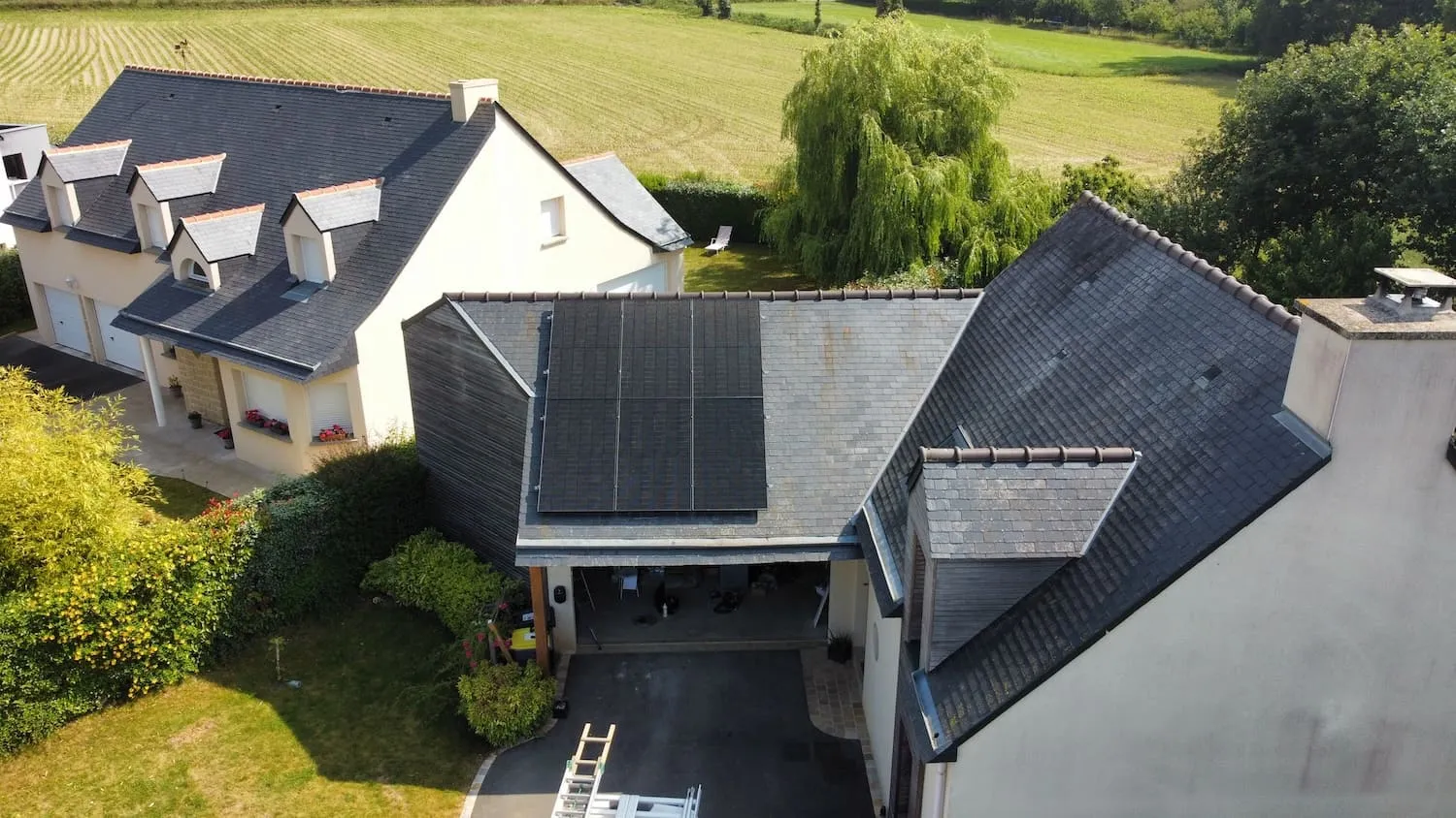 Aerial view of a suburban house with six solar panels installed on a roof section, surrounded by greenery and fields.