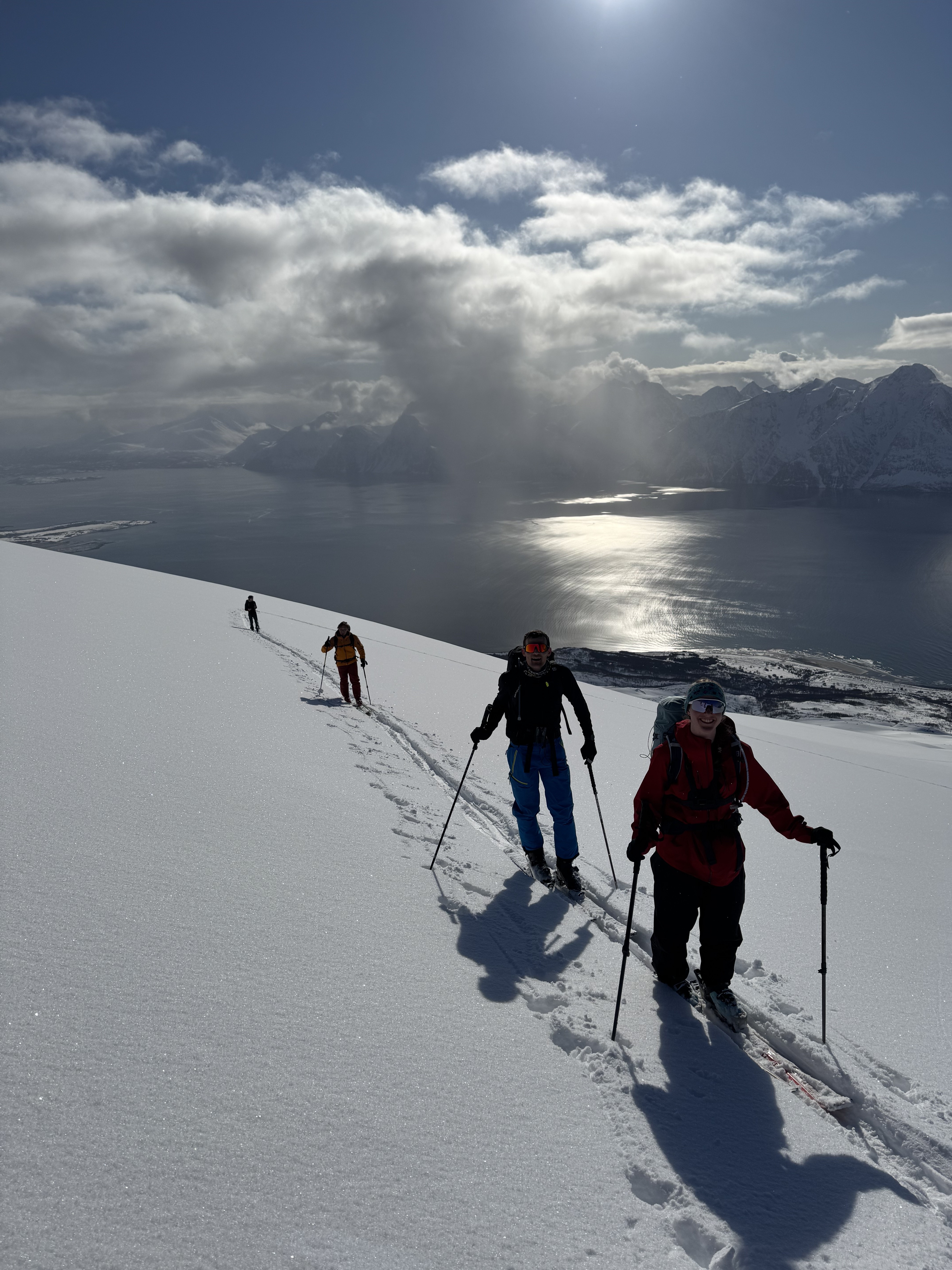 Four people ski touring uphill on a snowy mountain slope with a fjord and cloud-covered mountains in the background under a sunny sky.