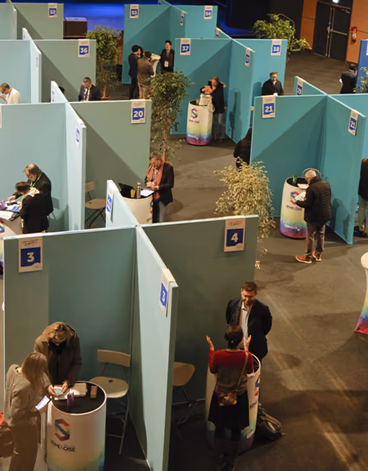 Vue en plongée d'un espace d'exposition ou de foire avec des stands numérotés disposés en rangées, séparés par des cloisons bleues. Plusieurs personnes discutent ou consultent des documents aux stands, certains avec des comptoirs arrondis portant un logo coloré. Des plantes décoratives sont placées entre les stands.