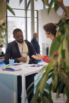 Deux personnes assises à une table dans un bureau moderne, en train de discuter, avec des plantes vertes en avant-plan et une grande fenêtre en arrière-plan.