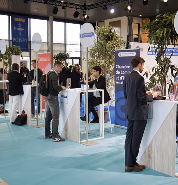 Vue intérieure d'un salon professionnel ou d'une foire commerciale avec plusieurs stands où des personnes discutent, dont un homme en costume debout près d'un comptoir blanc, et un homme en vêtements décontractés parlant avec une femme assise, avec des plantes et des bannières publicitaires en arrière-plan.