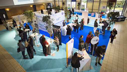 Vue aérienne d'un hall d'exposition avec plusieurs stands d'entreprise, des visiteurs discutant et consultant des documents, et des plantes décoratives au centre.