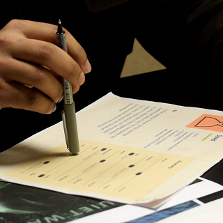 Hand holding a pen pointing to a filled multiple-choice answer sheet on a desk with documents.