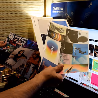 Person holding a fan of colorful printed images and photographs in front of a computer screen displaying various images.