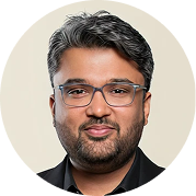 Portrait of a man with glasses, dark hair, and a trimmed beard, wearing a black jacket and navy shirt, smiling slightly on a light background.
