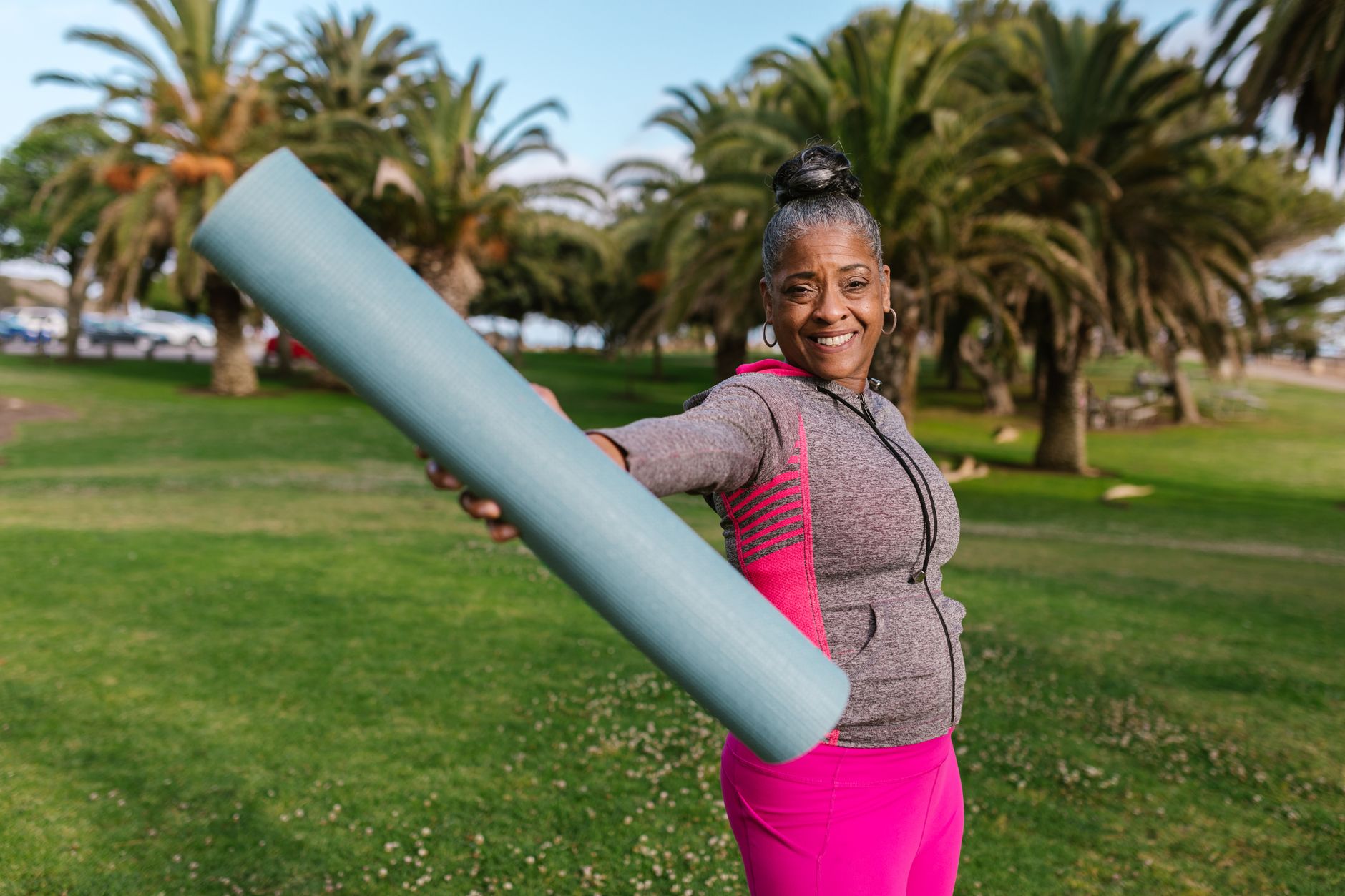 Smiling senior woman holding yoga mat, preparing for outdoor exercise in a sunny park — Longevity & Healthspan