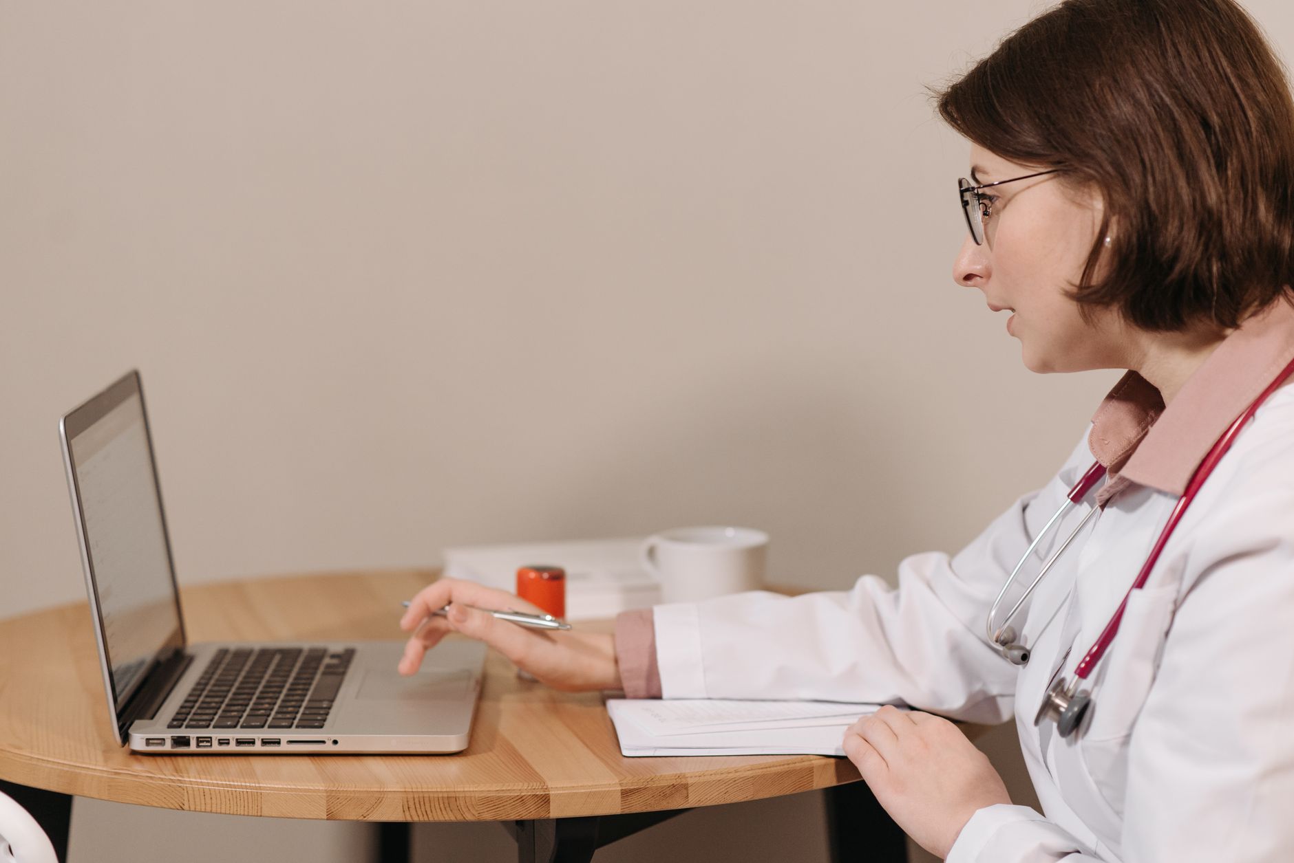 A female doctor in a white coat using a laptop while writing notes at a wooden table — AI & Digital Health