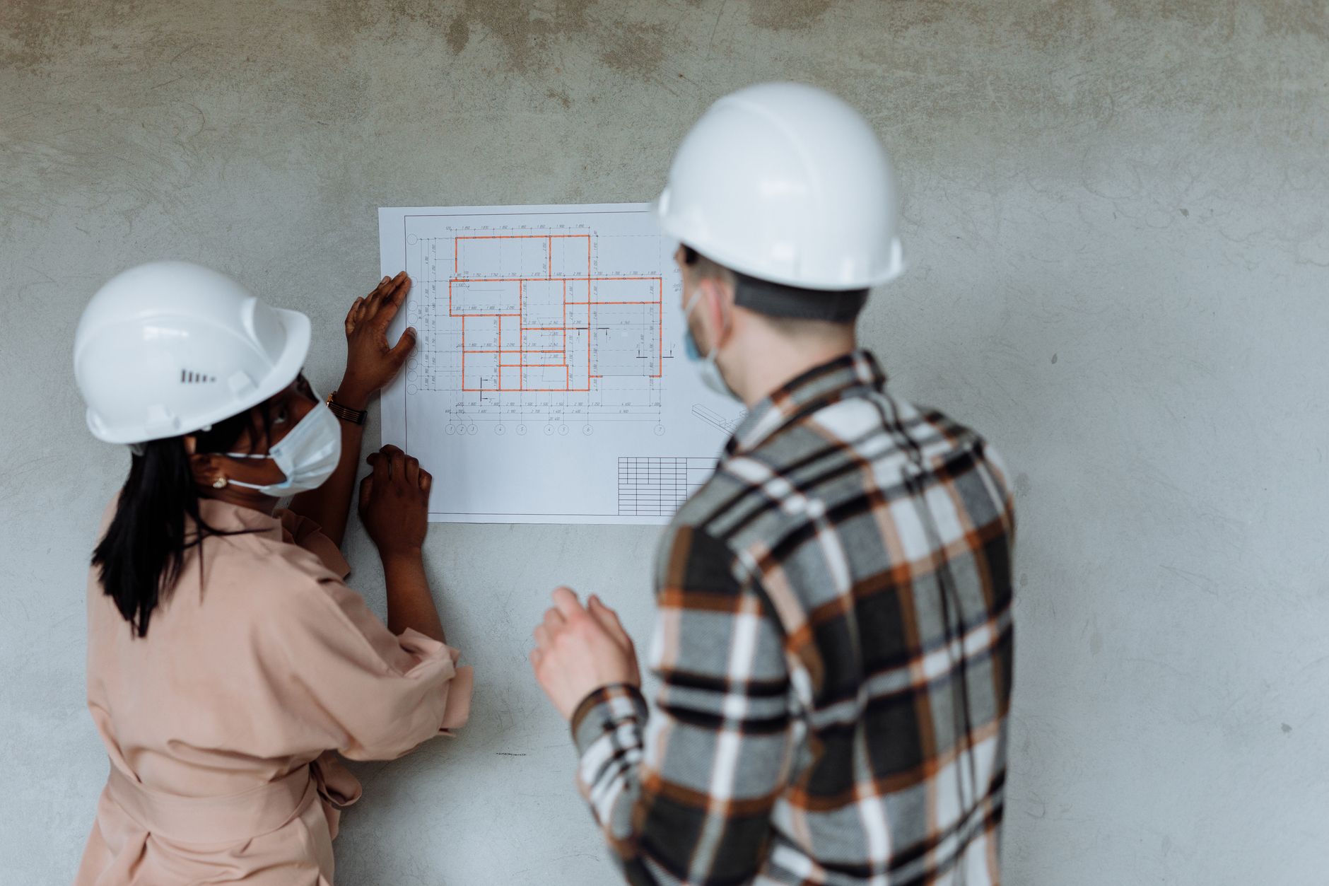 Two architects in hard hats and masks review blueprints during a building project — Healthcare Infrastructure