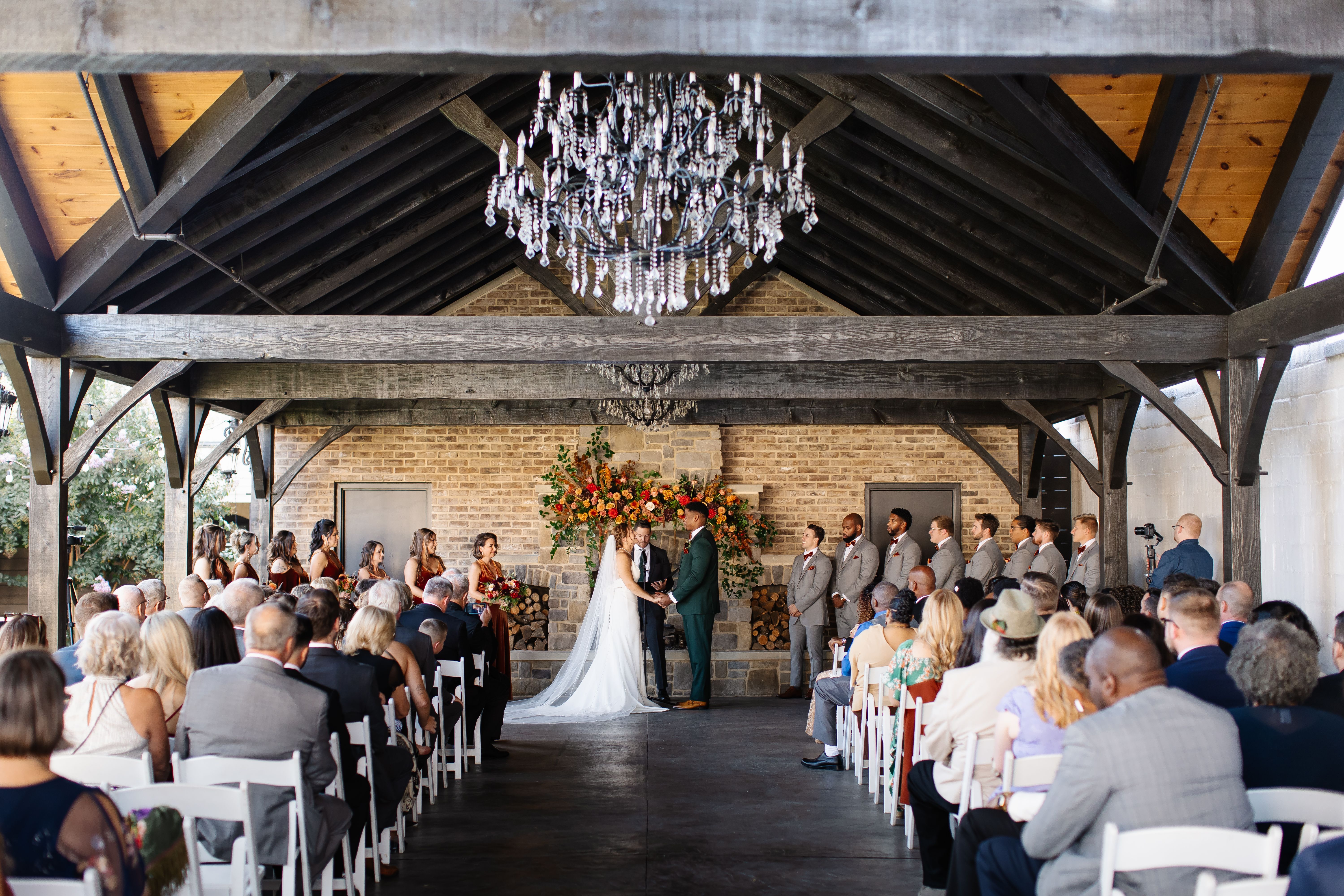 Bride and groom exchanging vows during an indoor wedding ceremony at The Bond, a historic wedding venue in Downtown York, Pennsylvania, featuring exposed beams, crystal chandeliers, and a stone fireplace.